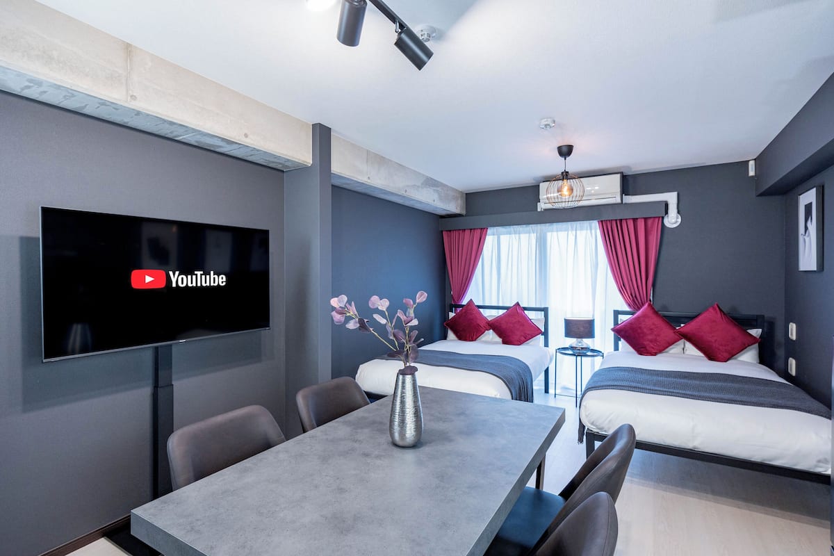 Une chambre d’hôtel moderne équipée d’un téléviseur à écran plat affichant le logo de YouTube, d’une table à manger agrémentée d’un vase de fleurs, de deux lits avec des oreillers rouges et d’une grande fenêtre avec des rideaux.