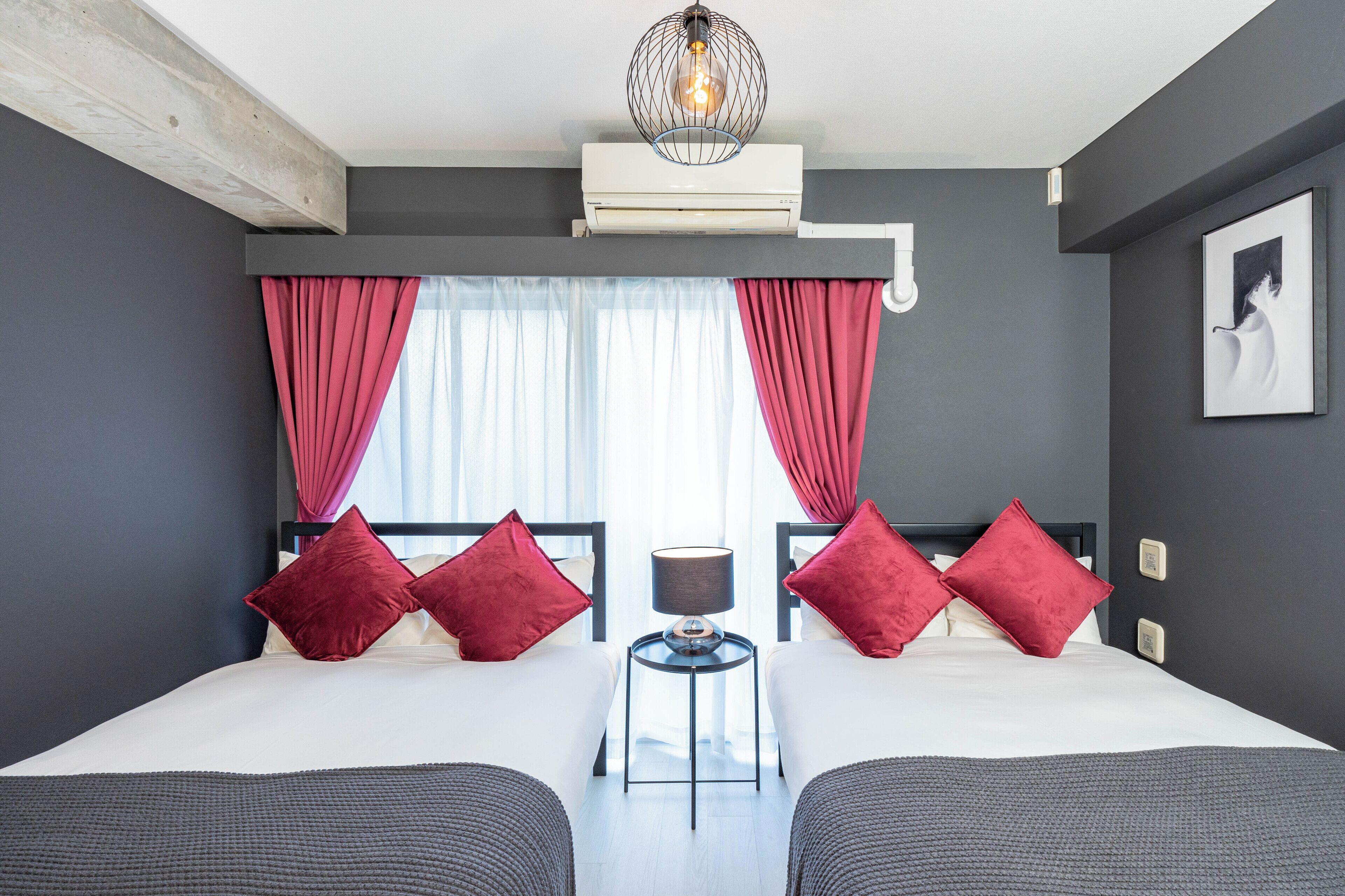 A modern hotel room with a large bed, two red pillows, a small table with a lamp, and a view of the outdoors through sheer curtains.
