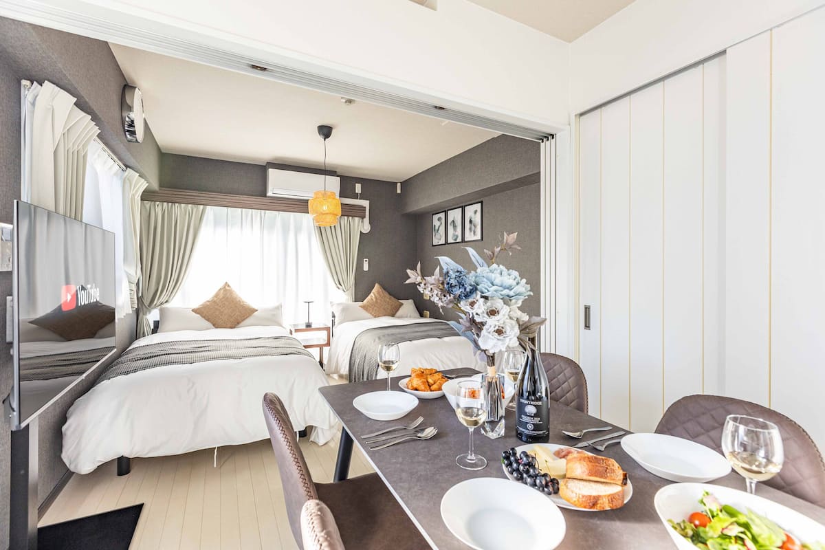 Une chambre d’hôtel moderne avec un lit, une table à manger dressée avec des assiettes, des verres et une bouteille de vin, ainsi qu’un vase à fleurs.