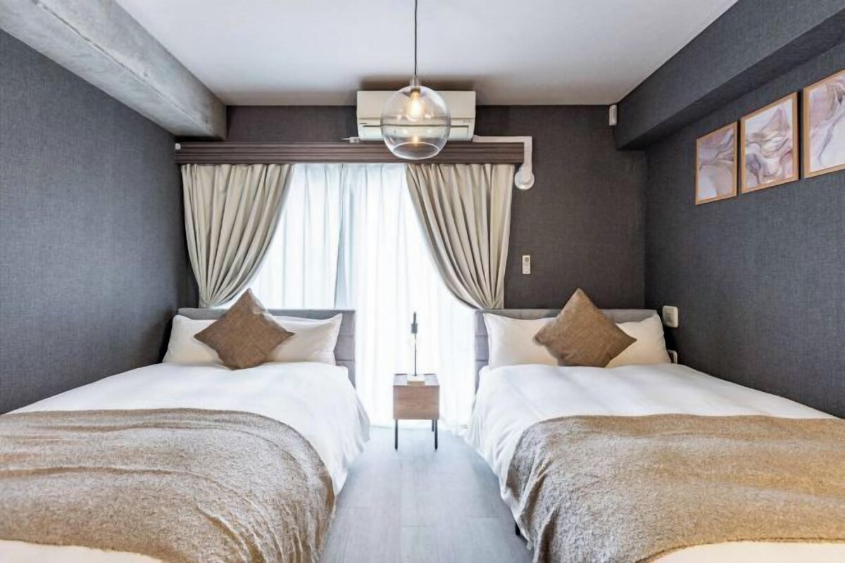 A hotel room with two beds, a bedside table, a hanging light fixture, and a window with curtains.