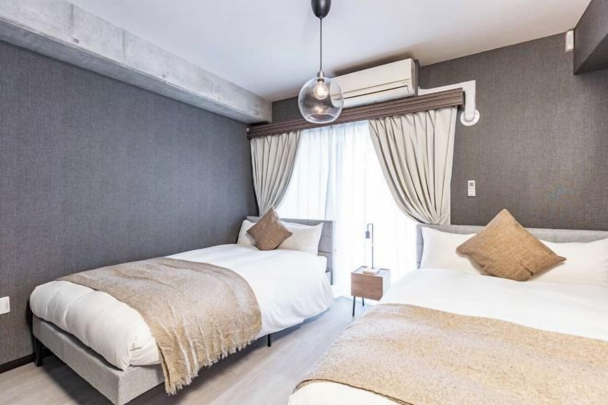 A modern hotel room with two beds, a large window with curtains, a hanging light fixture, and a small bedside table.