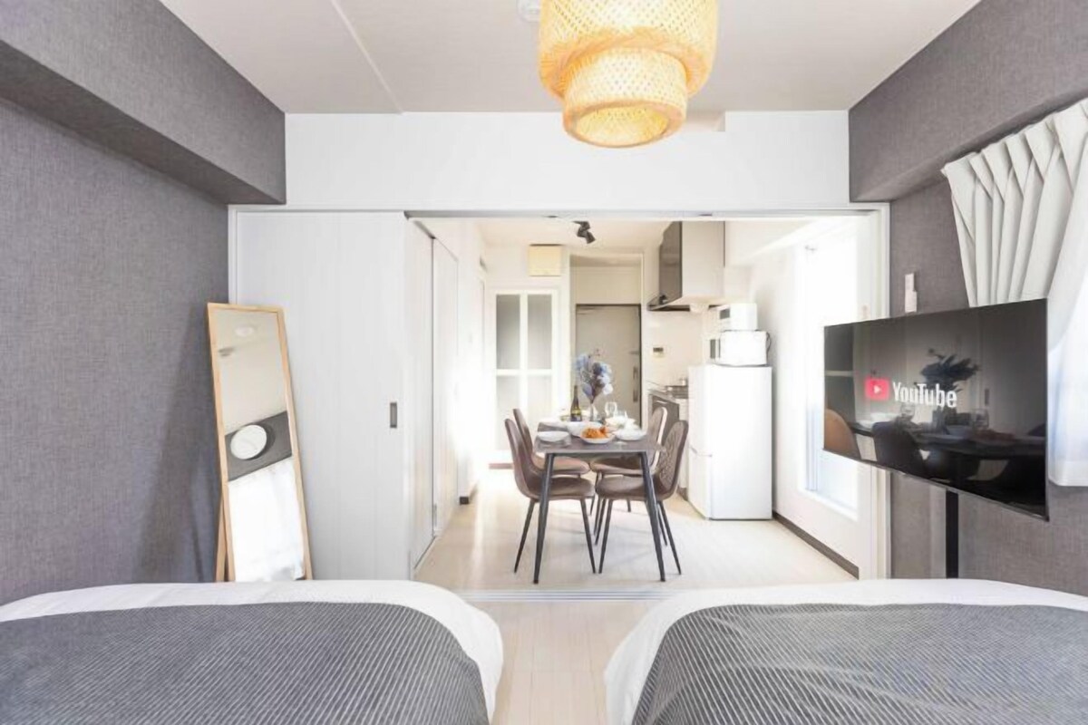 A hotel room with two beds, a dining table, a TV, and a kitchen area visible through an open door.