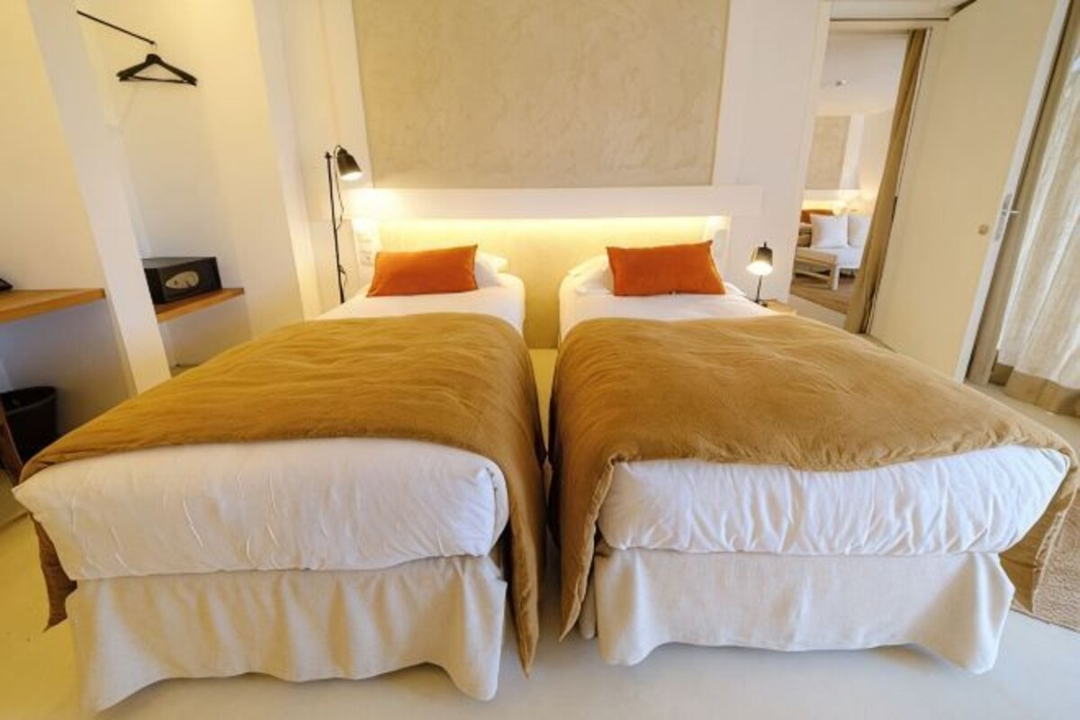 A double room with two beds, each with a white mattress and brown headboard, and a single bed with a white mattress and orange pillow.