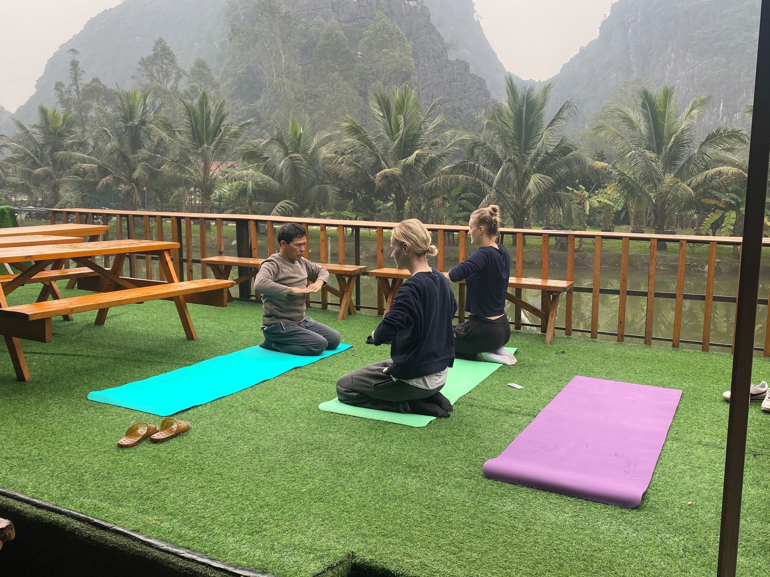 Un cours de yoga donné sur une terrasse enherbée, dotée de bancs en bois et de tables de pique-nique, avec vue sur un paysage montagneux parsemé de palmiers.