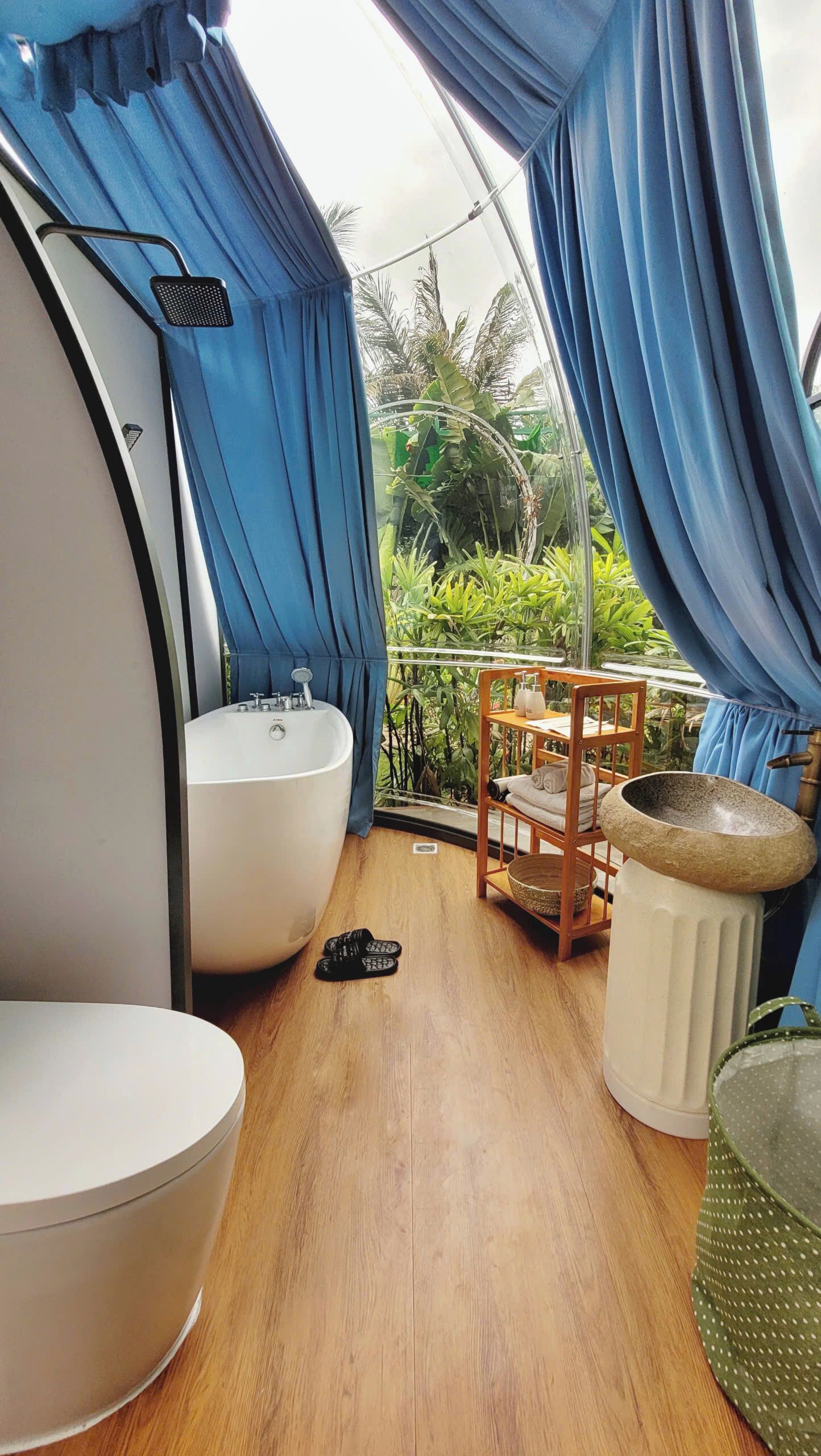 A bathroom with a bathtub, sink, and shower, wooden flooring, and a view of greenery outside.