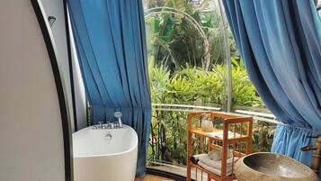 A bathroom with a bathtub, sink, and shower, wooden flooring, and a view of greenery outside.