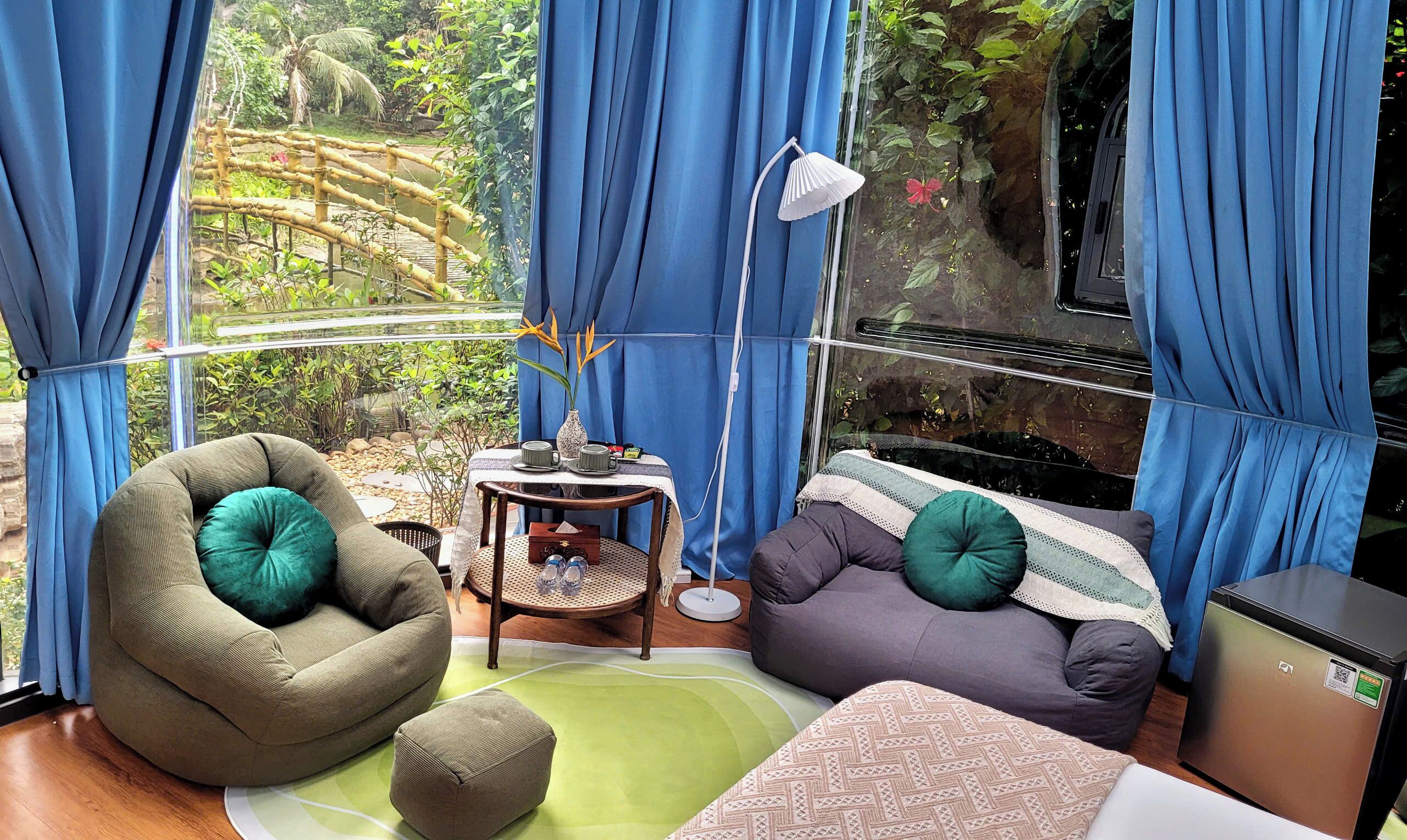 Habitación con vista a un puente de bambú y follaje, con un sillón de mimbre verde, un sofá gris con un cojín verde, una mesita redonda con una lámpara y un refrigerador compacto.