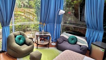 Habitación con vista a un puente de bambú y follaje, con un sillón de mimbre verde, un sofá gris con un cojín verde, una mesita redonda con una lámpara y un refrigerador compacto.