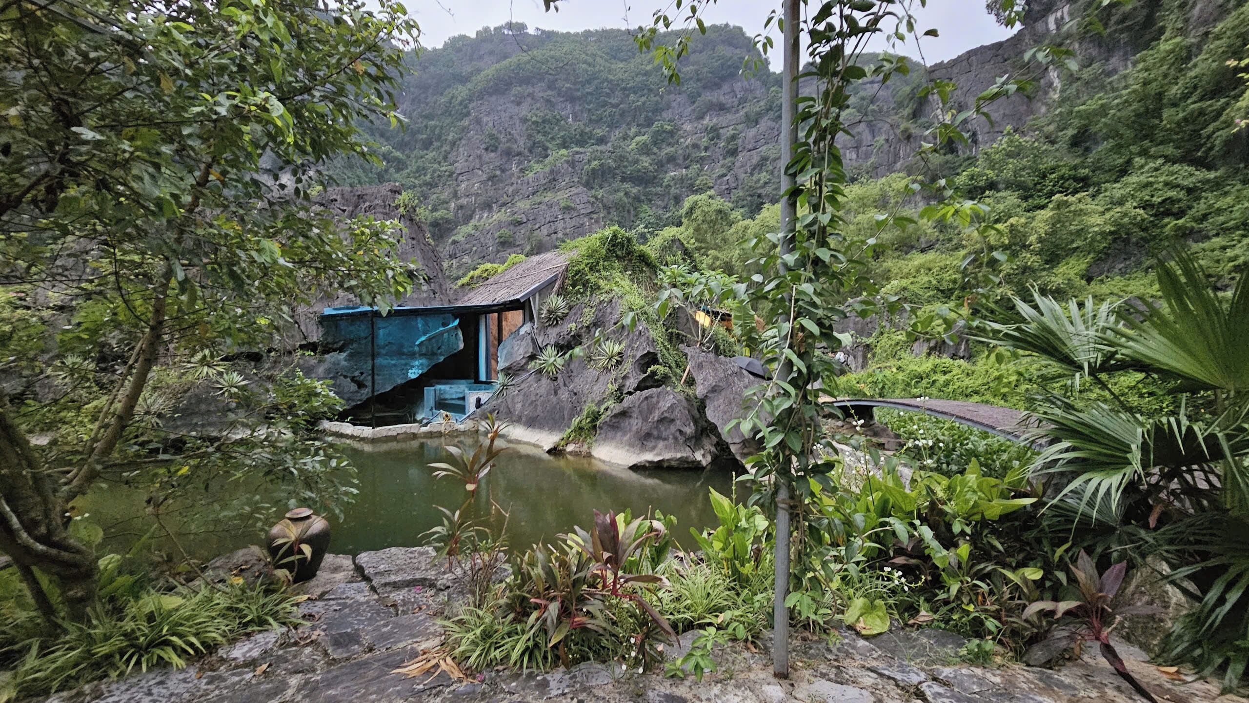 A secluded cabin with a pond, surrounded by lush greenery and rocky terrain.