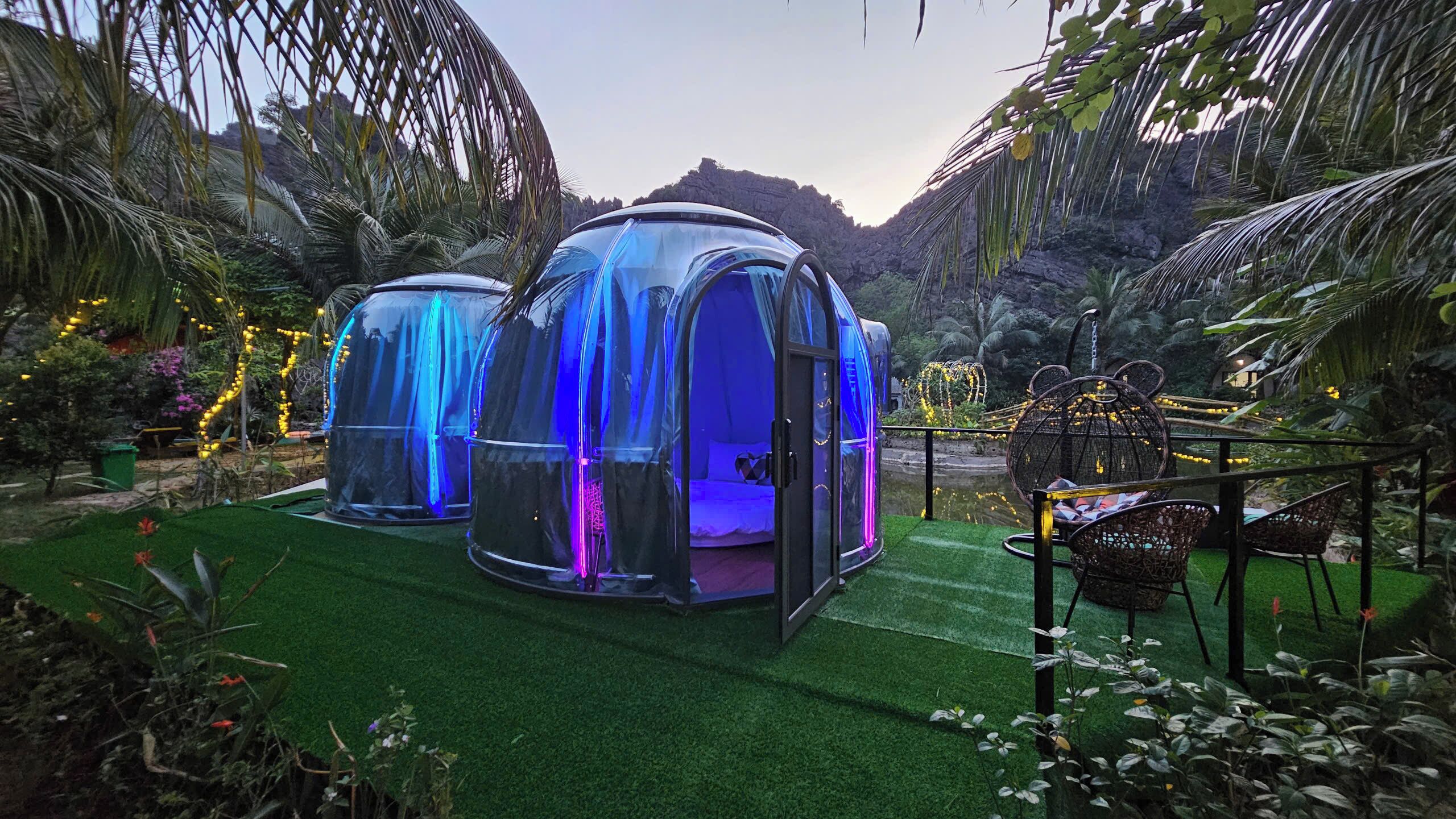 Two transparent dome-shaped structures with a bed inside, surrounded by lush greenery and decorative lights.