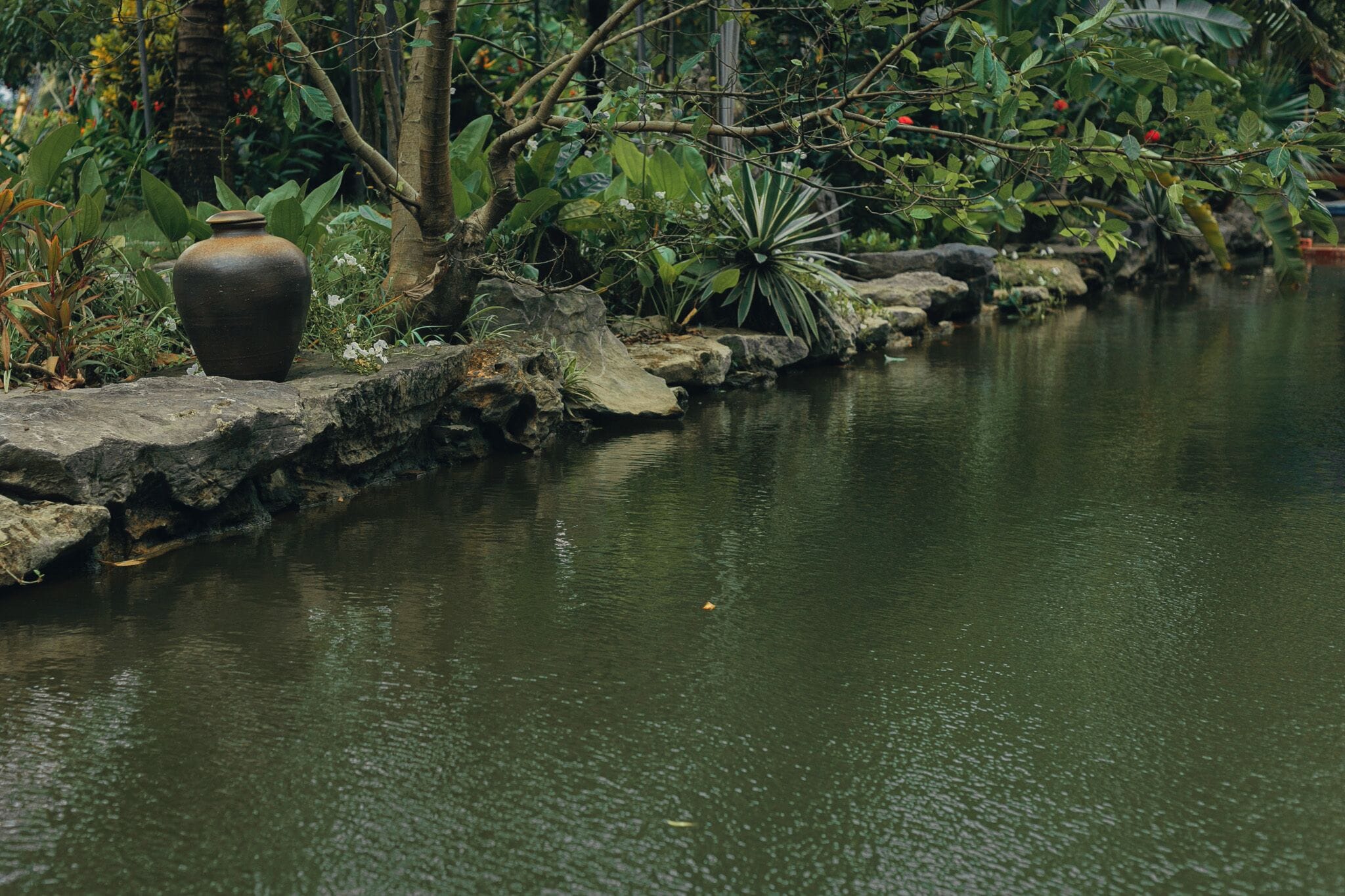Um jardim tranquilo com um vaso grande sobre uma rocha, perto de um lago calmo.