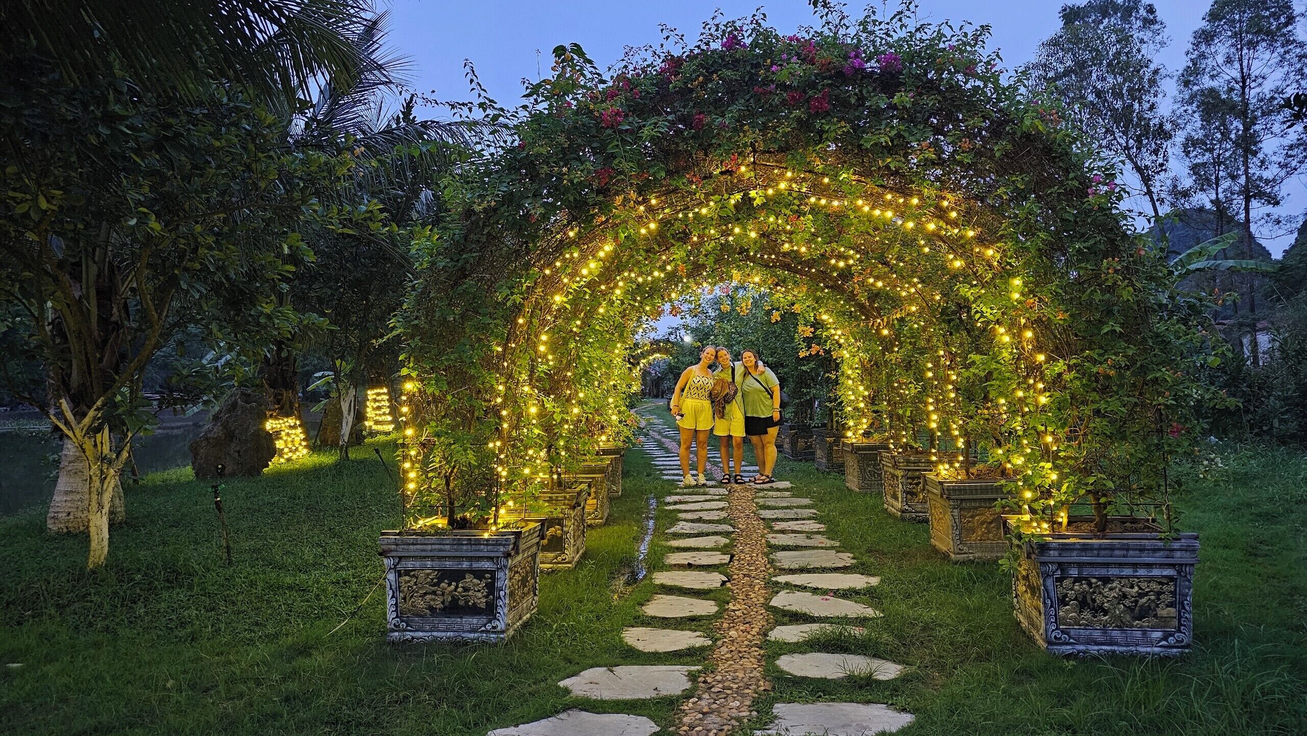 An outdoor garden pathway with an archway decorated with lights, surrounded by greenery and stone planters.