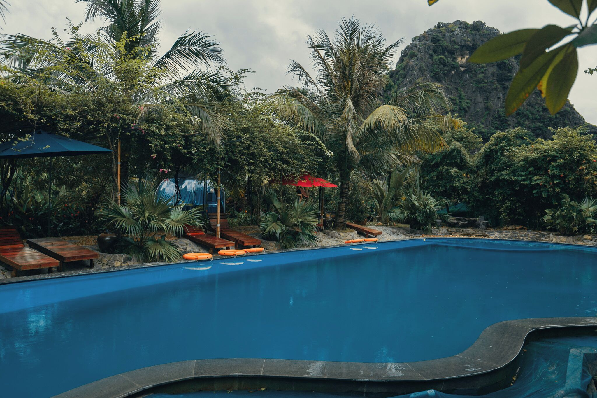 A swimming pool with wooden loungers and orange life vests, surrounded by lush tropical vegetation and a mountain in the background.