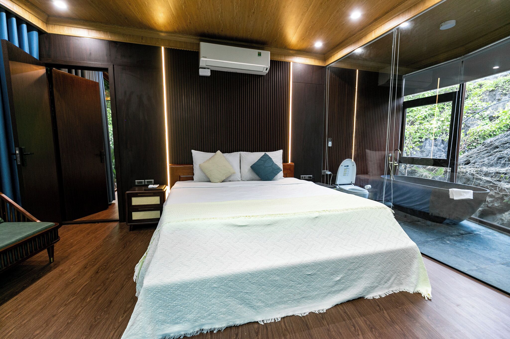 A modern bedroom with a large bed, wooden flooring, a built-in bench, and a view of a rocky landscape through a glass door.