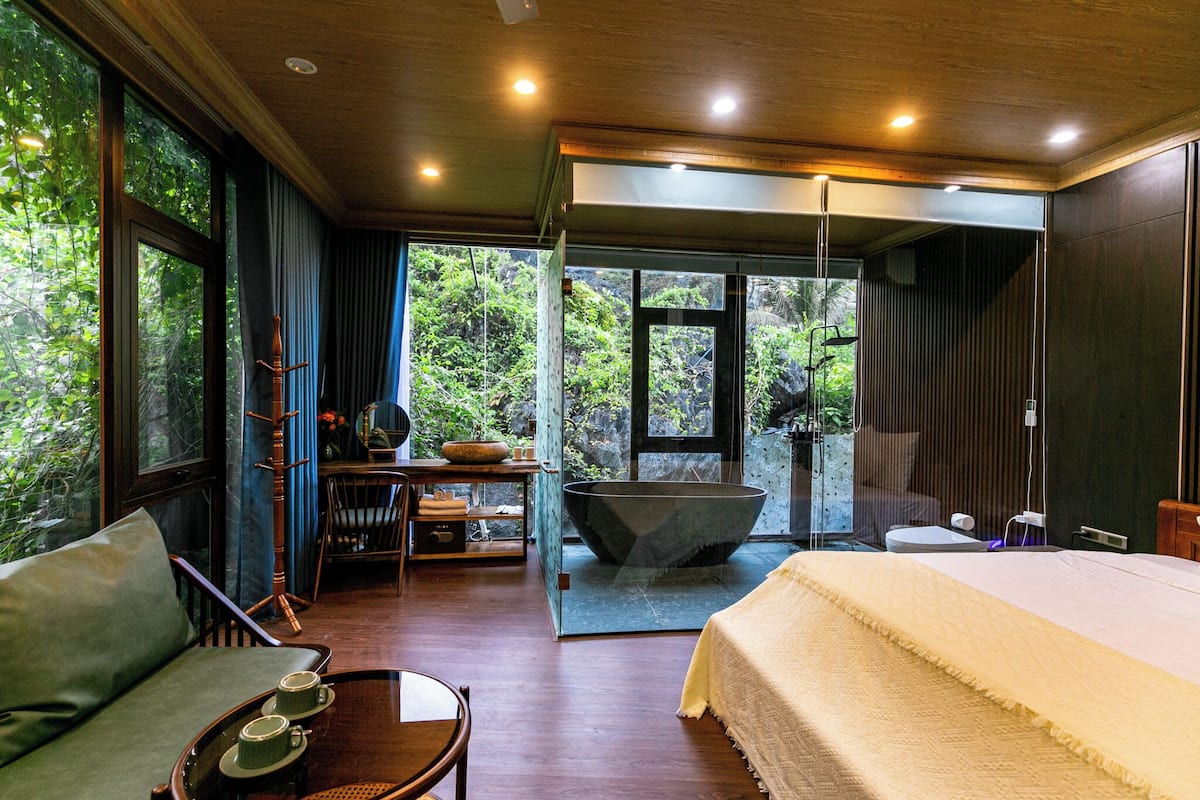 Un dormitorio moderno con una cama grande, una zona de estar con una mesa y sillas, una bañera y vistas a vegetación exuberante a través de amplios ventanales.
