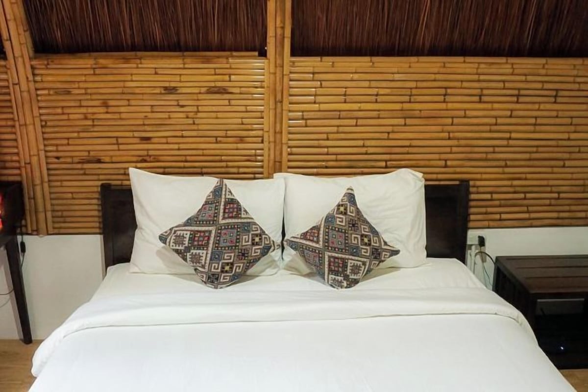 Uma cama com roupa de cama branca e almofadas estampadas, uma cabeceira de madeira e uma mesa de cabeceira com luminária.