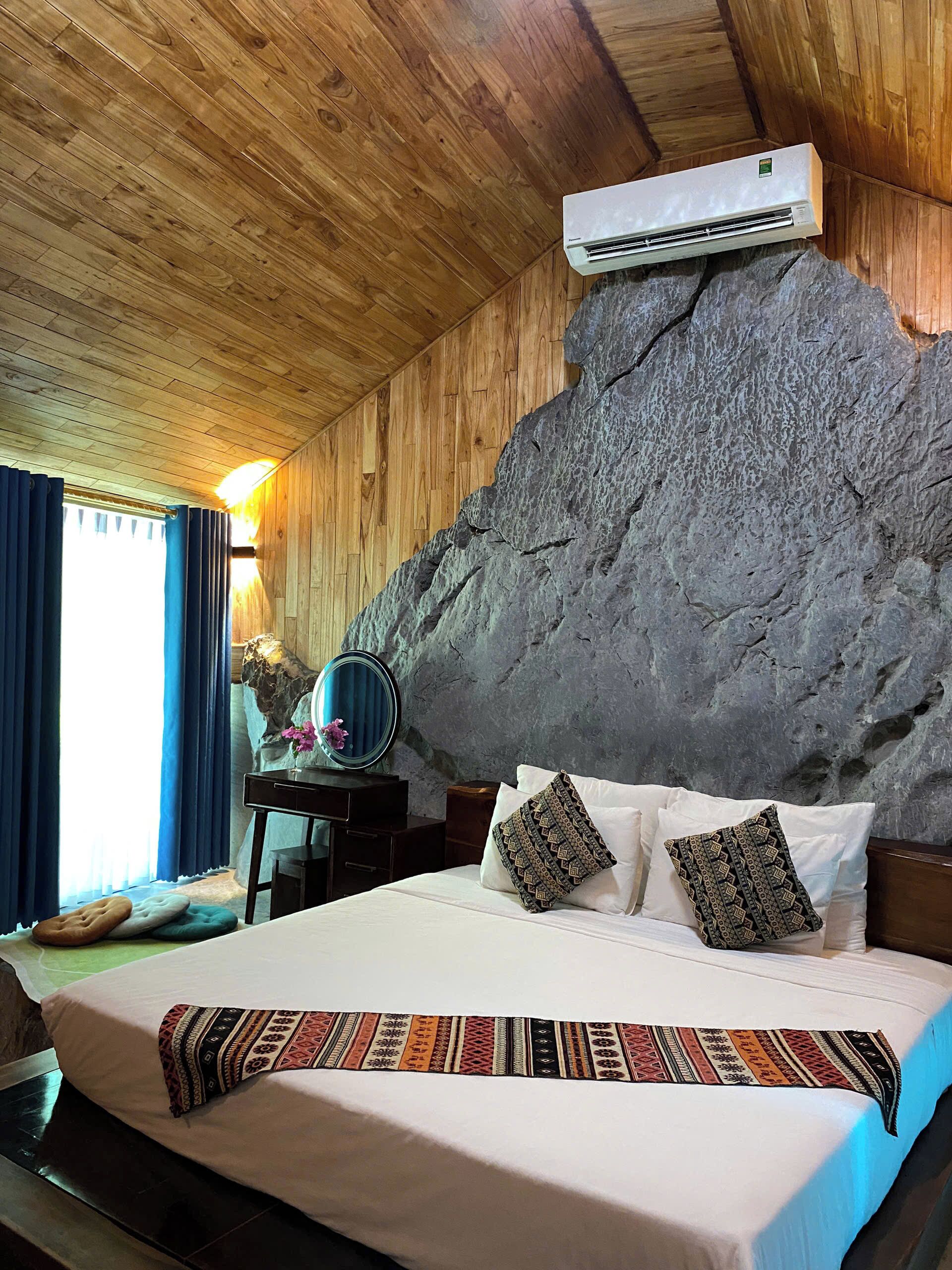 Quarto com teto de madeira, parede de pedra, cama com almofadas estampadas, mesa de cabeceira com espelho e ar condicionado.