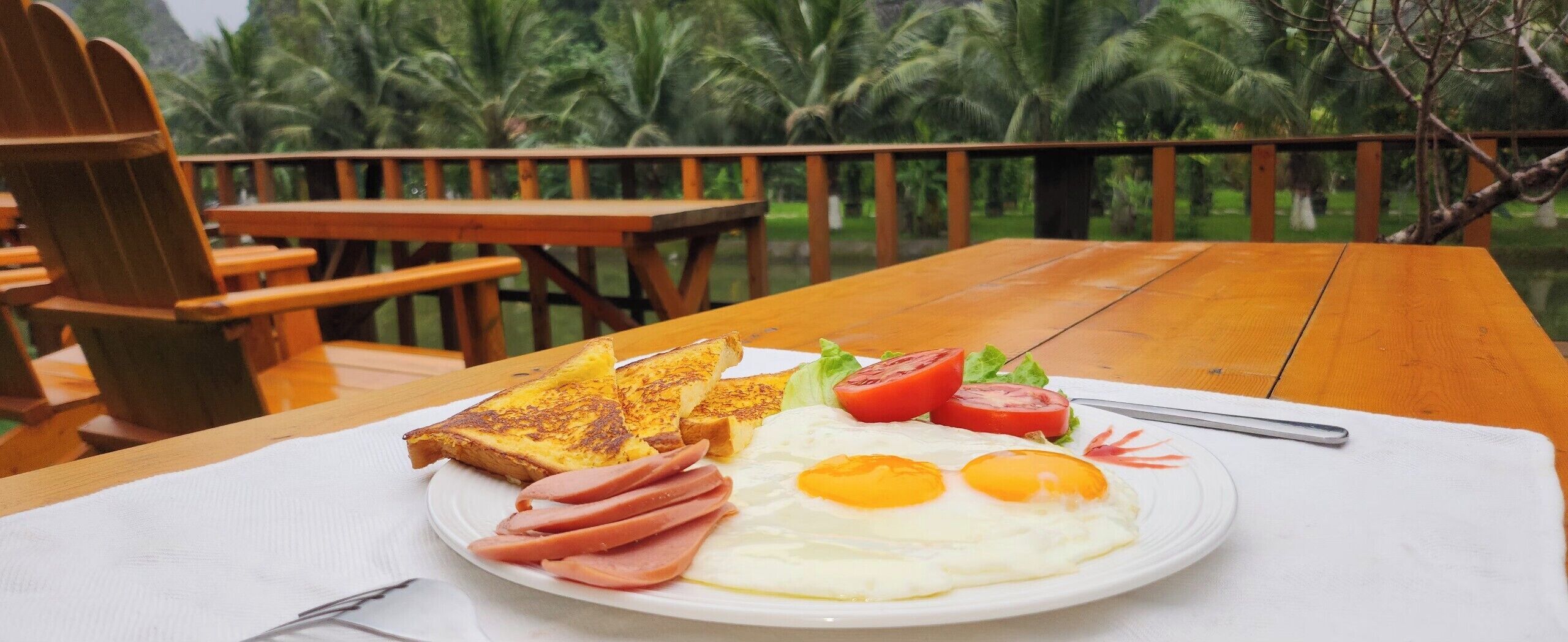 Un plato de desayuno con tostadas, huevos, jamón y tomates sobre una mesa de madera al aire libre.