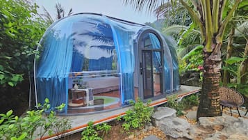 Una estructura abovedada transparente con una cama y zona de estar en su interior, rodeada de follaje y rocas.