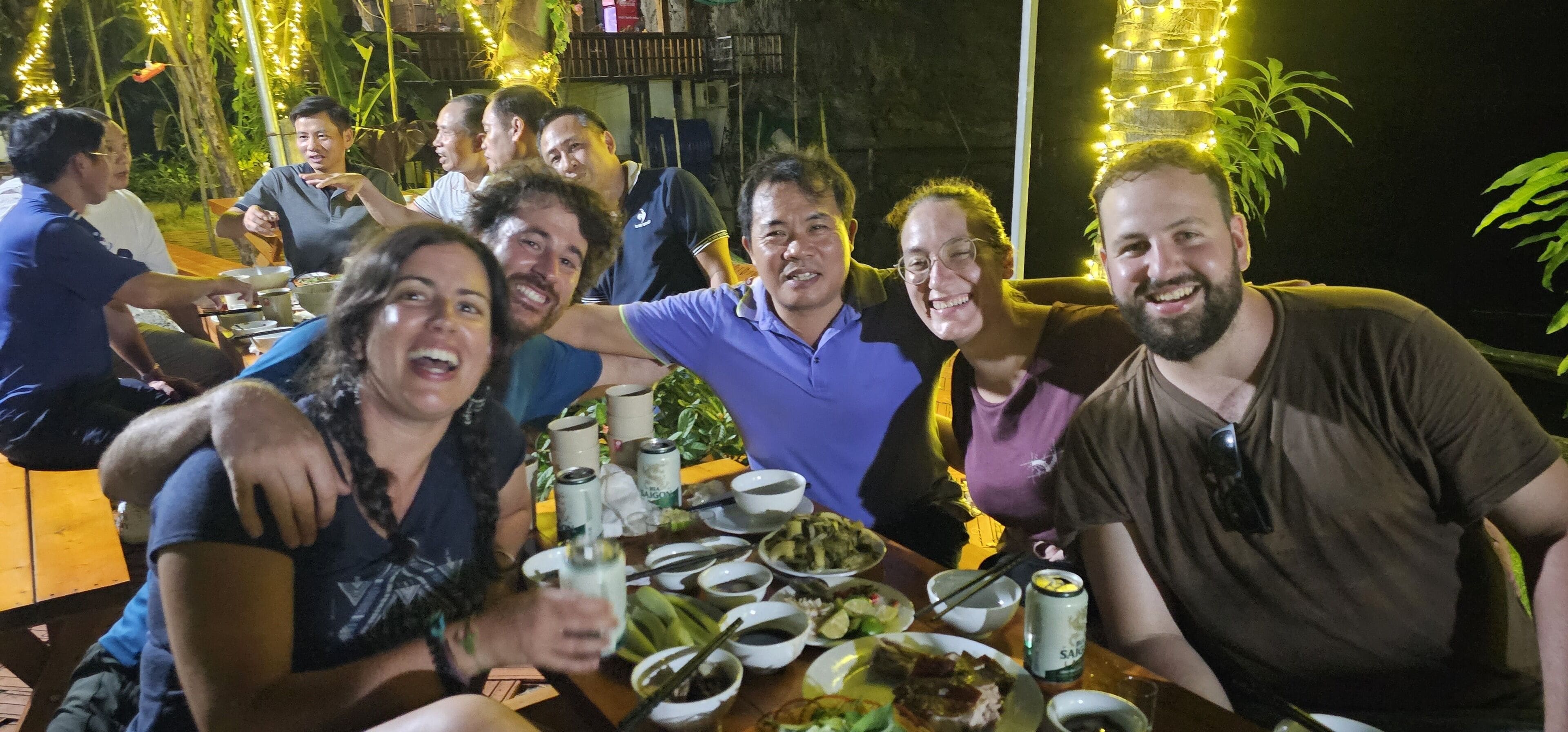A group of people dining outdoors at night with string lights and plants in the background.
