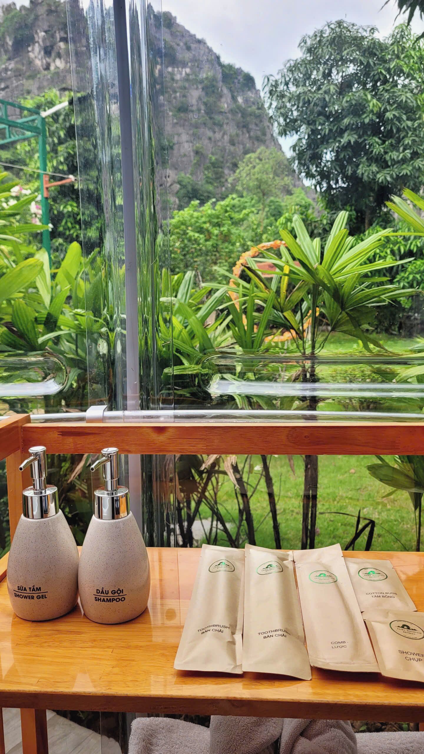 A wooden table with two shower gel bottles and four toiletry bags in a room with a view of a waterfall and greenery.