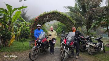 Three people standing in front of motorcycles, with a lush garden and a decorative archway in the background.