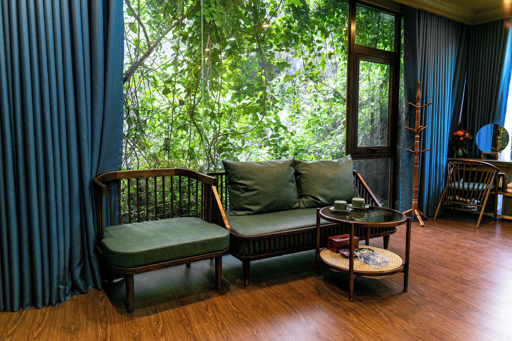 A room with a wooden floor, a sofa with cushions, a small round table, and a chair. There is a window with blue curtains, and a wooden coat rack is visible.