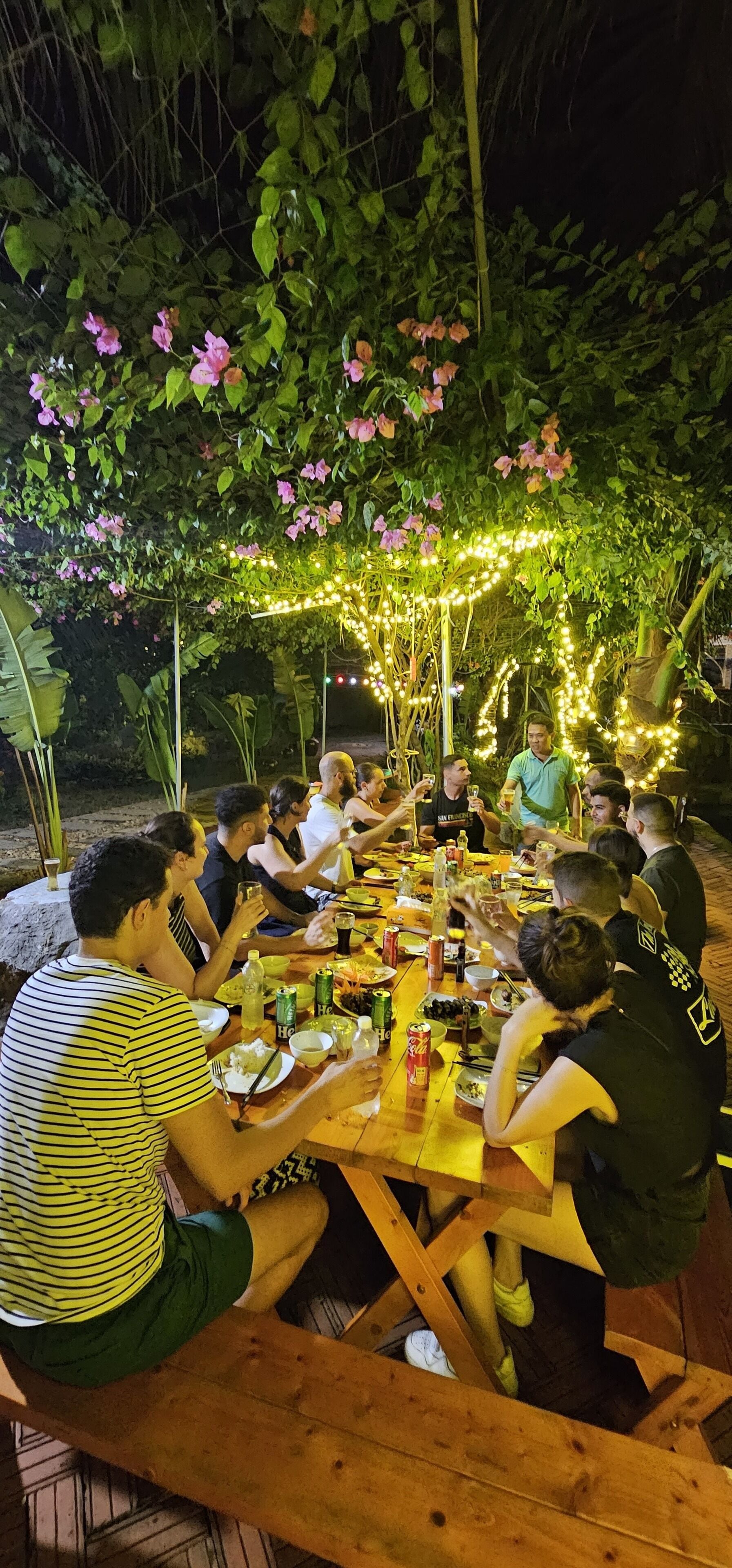 A group of people dining outdoors at a wooden table under a canopy of flowering vines and string lights.