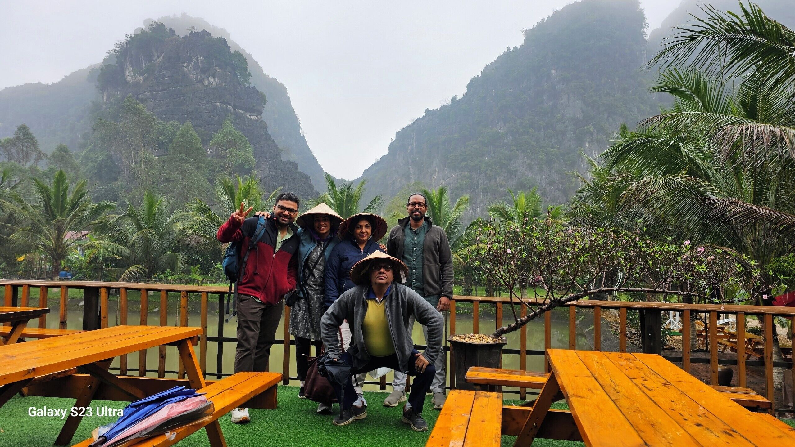 Un gruppo di persone che posa su un molo di legno con vista su un paesaggio montuoso e palme.