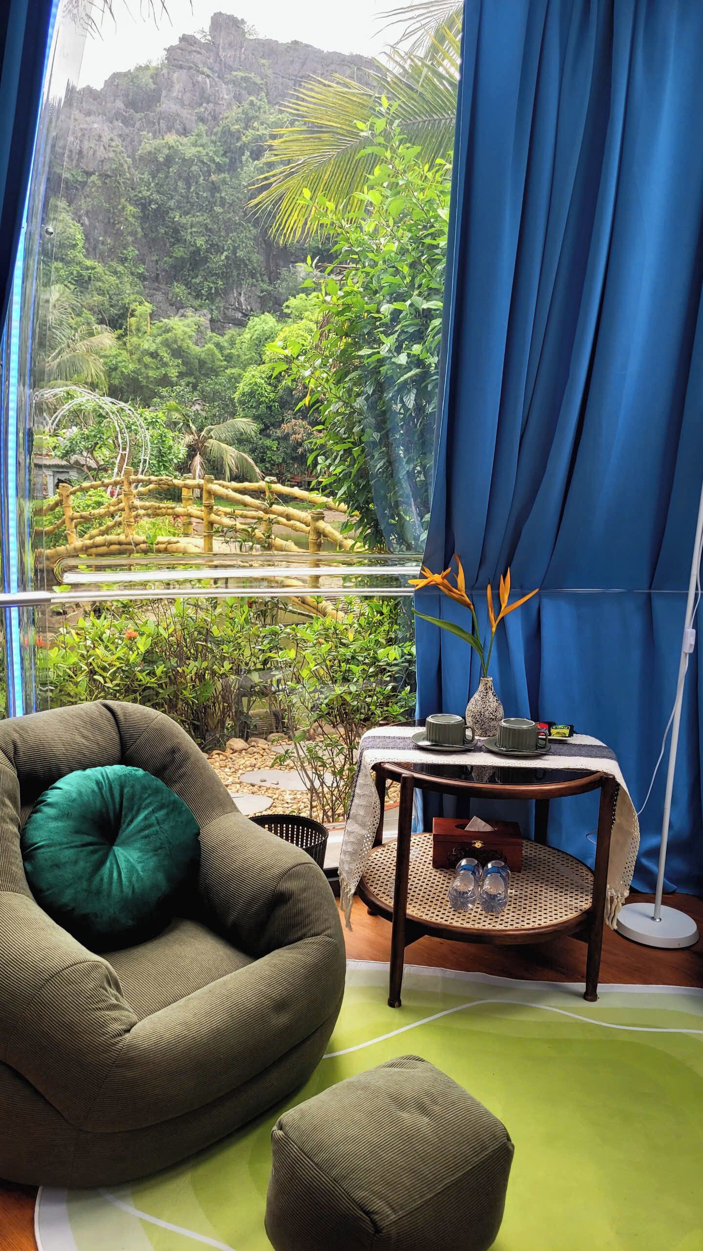 Ein Zimmer mit Blick auf einen üppigen Garten, ein grüner Sitzsack, ein kleiner runder Tisch mit einer Vase und einer Tasse sowie eine Stehlampe.