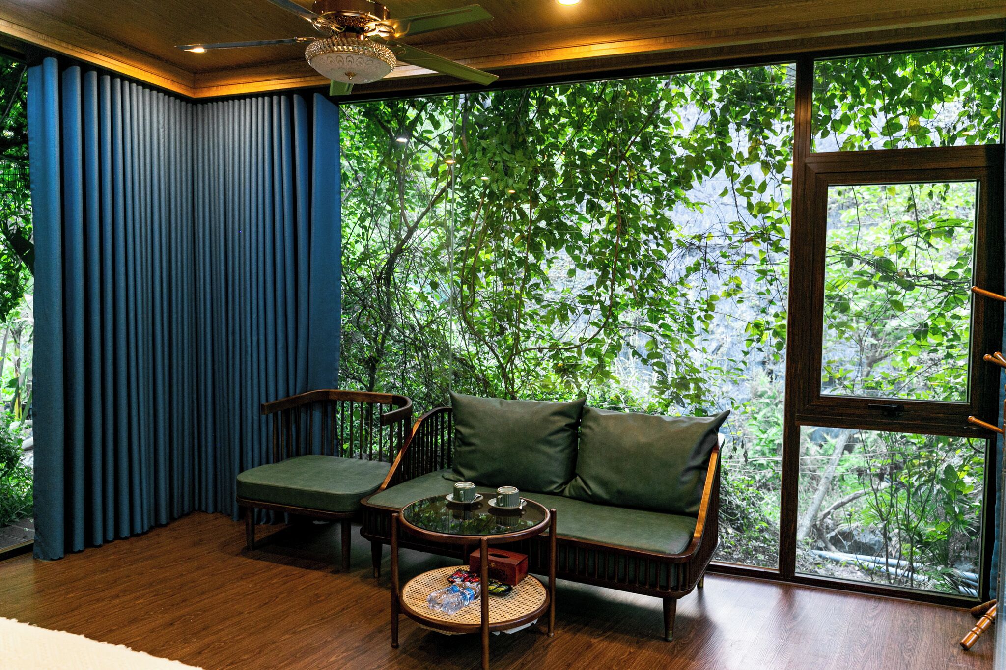 A room with a wooden floor, a green sofa, a round table with a glass top, and a view of trees through large windows.