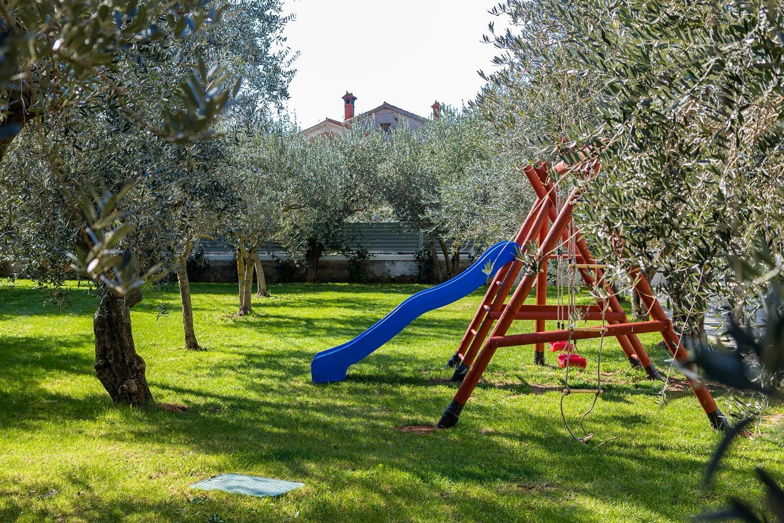 Un parc infantil amb tobogan blau i cadiretes en una zona verda plena d'oliveres.