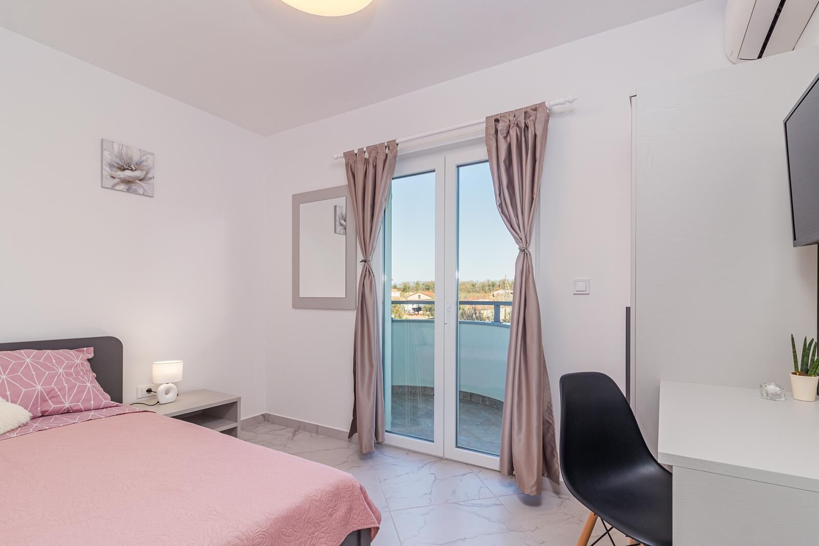 A bedroom with a bed, a desk, a chair, a TV, and a balcony with curtains.