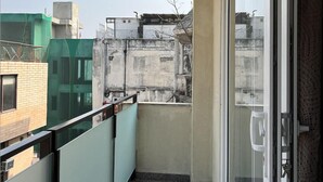 A balcony with a glass door, a metal railing, and a view of neighboring buildings. - Fully Furnished Room in Hauz Khas (New Delhi)