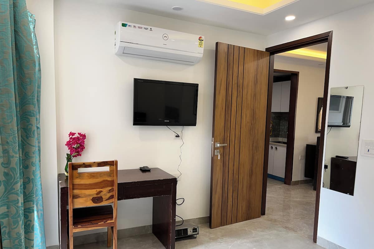 A room with a wooden desk, a chair, a TV mounted on the wall, and an air conditioning unit.