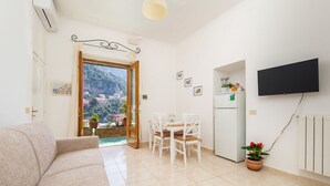 A living room with a sofa, a dining table with chairs, a TV mounted on the wall, a refrigerator, and a potted plant. - Rosa House - Breathtaking View of the Amalfi Coast (Positano)