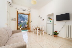 A living room with a sofa, a dining table with chairs, a TV mounted on the wall, a refrigerator, and a potted plant. - Rosa House - Breathtaking View of the Amalfi Coast (Positano)