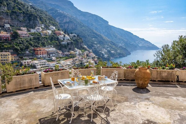 A terrace with a table set for breakfast, overlooking a coastal town with buildings on a hillside and the sea in the background. - Rosa House - Breathtaking View of the Amalfi Coast (Positano)