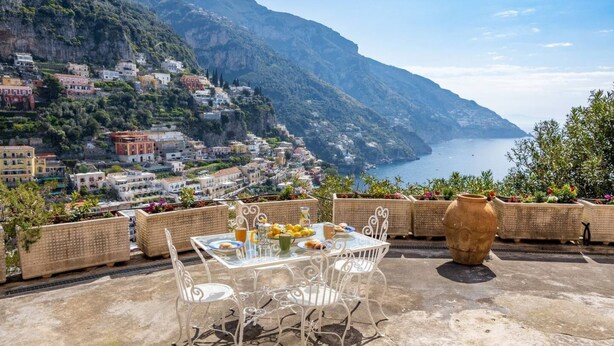 A terrace with a table set for breakfast, overlooking a coastal town with buildings on a hillside and the sea in the background. - Rosa House - Breathtaking View of the Amalfi Coast (Positano)