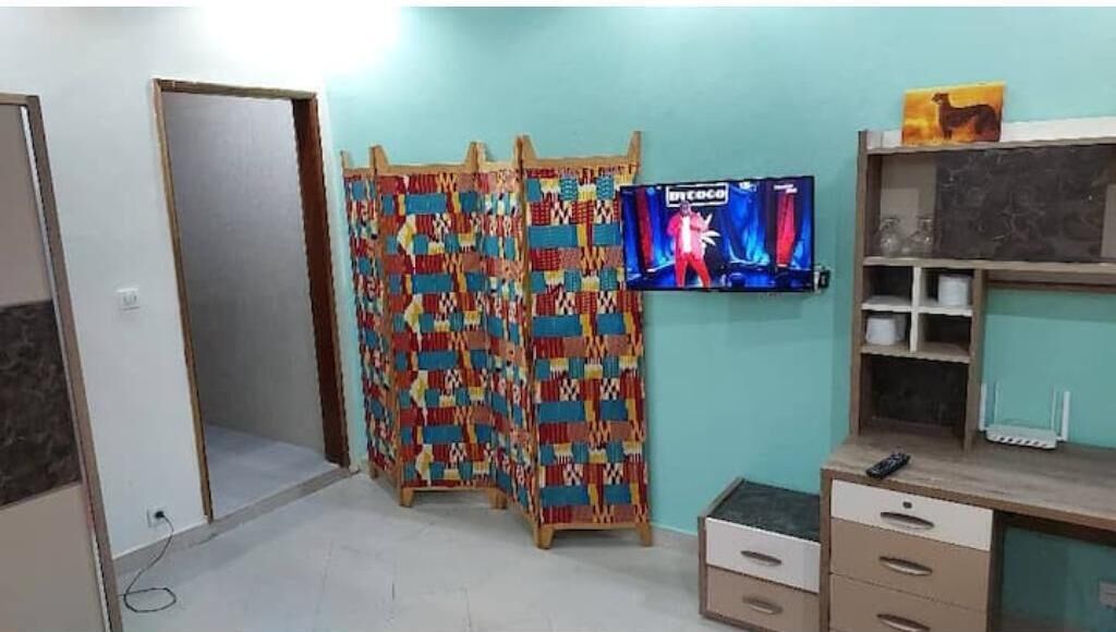 A room with a television on the wall, a patterned curtain, and a desk with drawers.