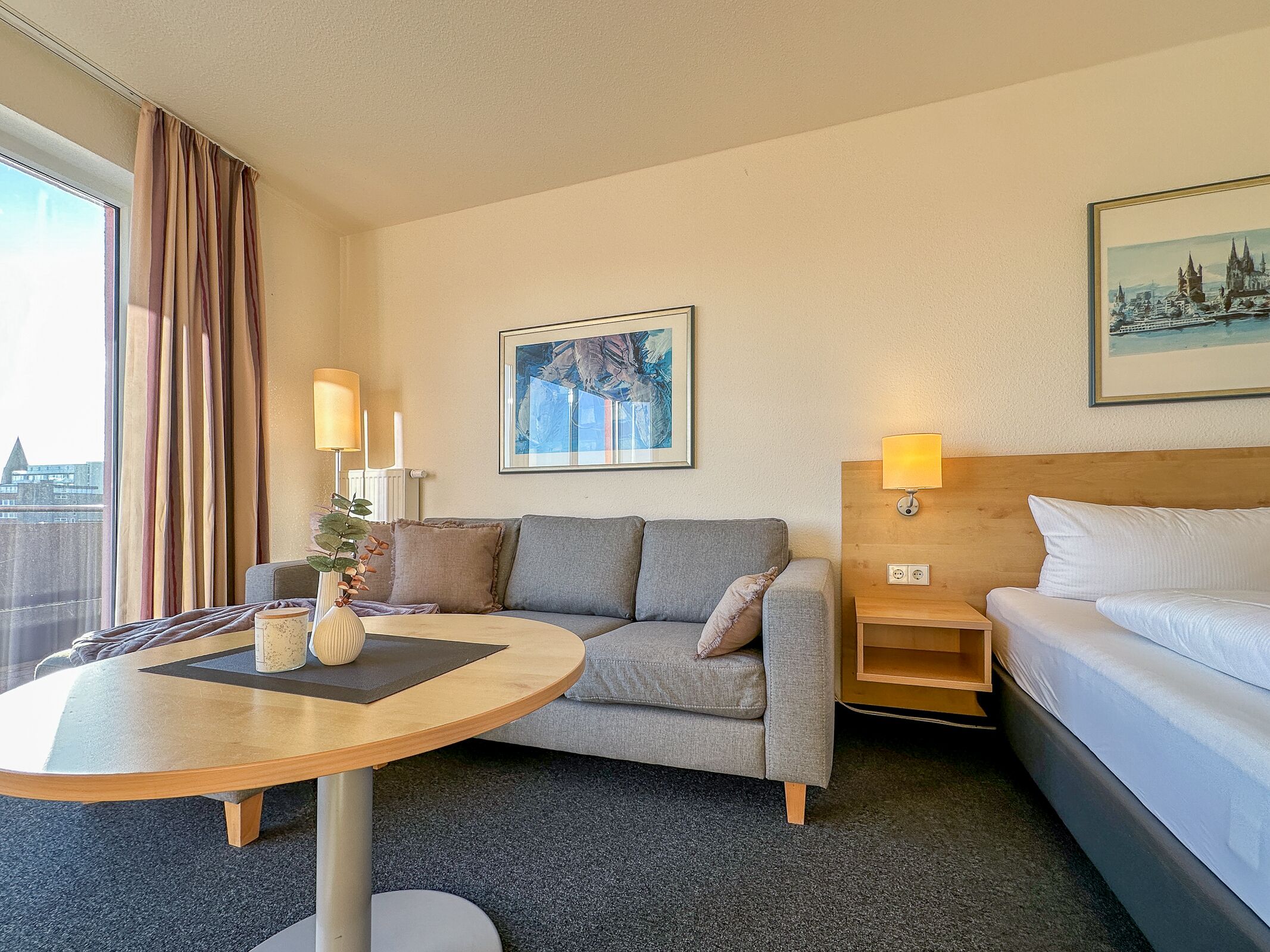 A hotel room with a grey sofa, a round wooden table, a bed, a nightstand with a lamp, and a painting on the wall.