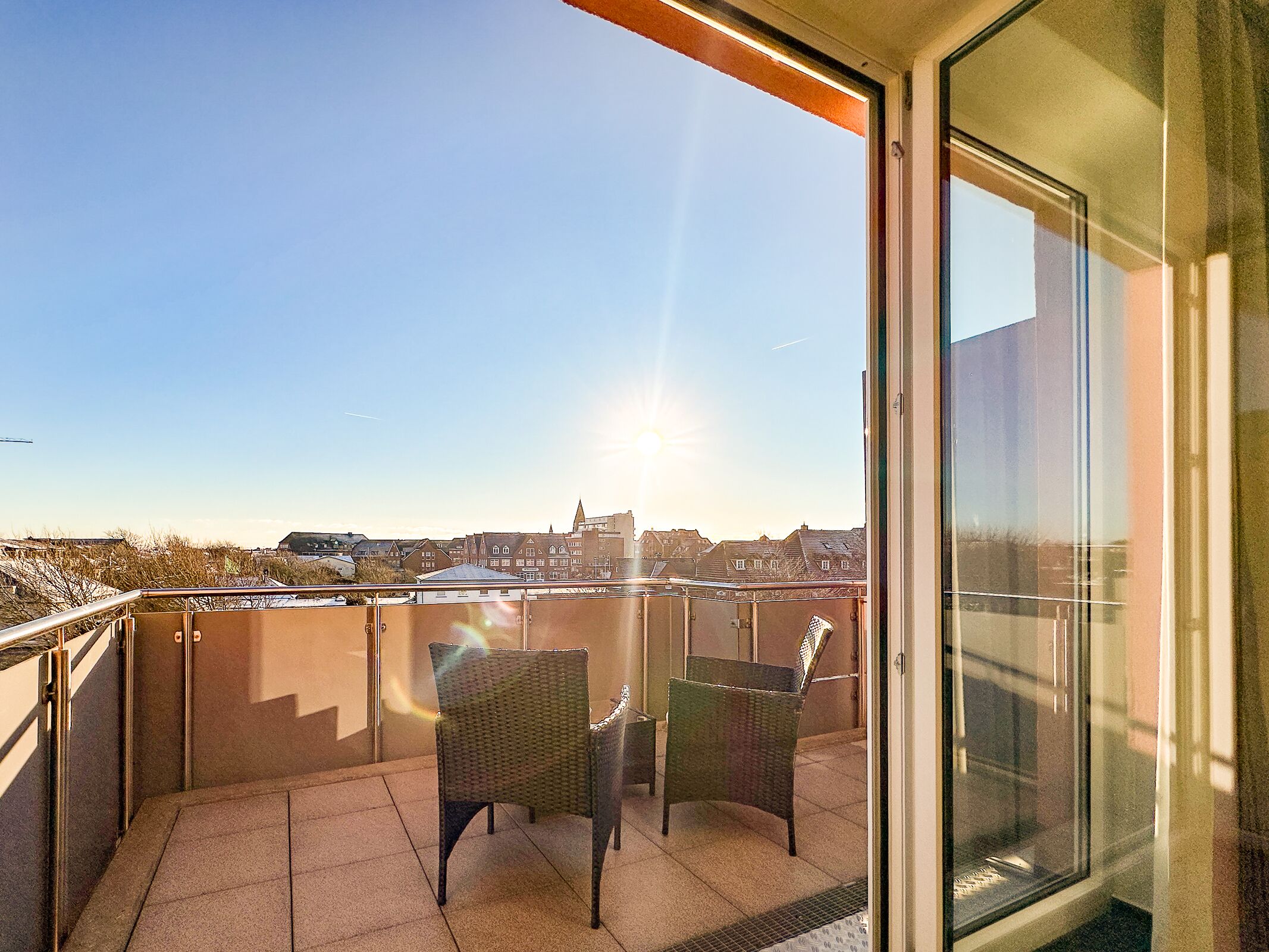 A balcony with two wicker chairs and a table, overlooking a cityscape with buildings and a clear sky.