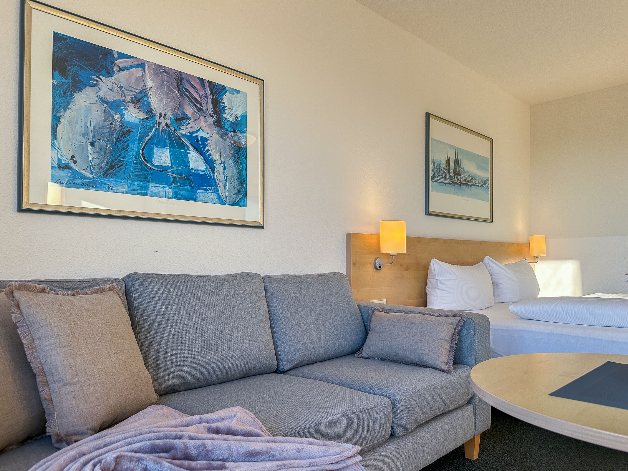 Ein Hotelzimmer mit grauem Sofa, rundem Couchtisch, Bett mit weißer Bettwäsche und zwei Wandlampen.
