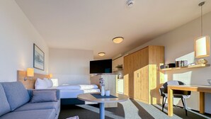 A compact hotel room with a sofa, a bed, a small round table, a TV, a wooden wardrobe, and a desk with a chair.