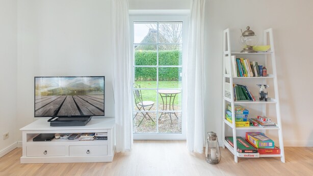 A living room with a TV on a white entertainment unit, a bookshelf with board games, and a view of a garden through a large window.