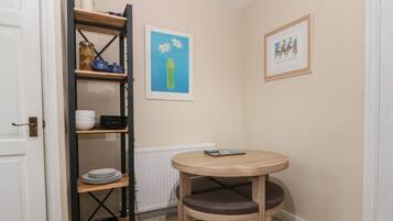 A compact kitchen with a round wooden table and a single chair, a black metal shelf unit with dishes, and two framed artworks on the wall.
