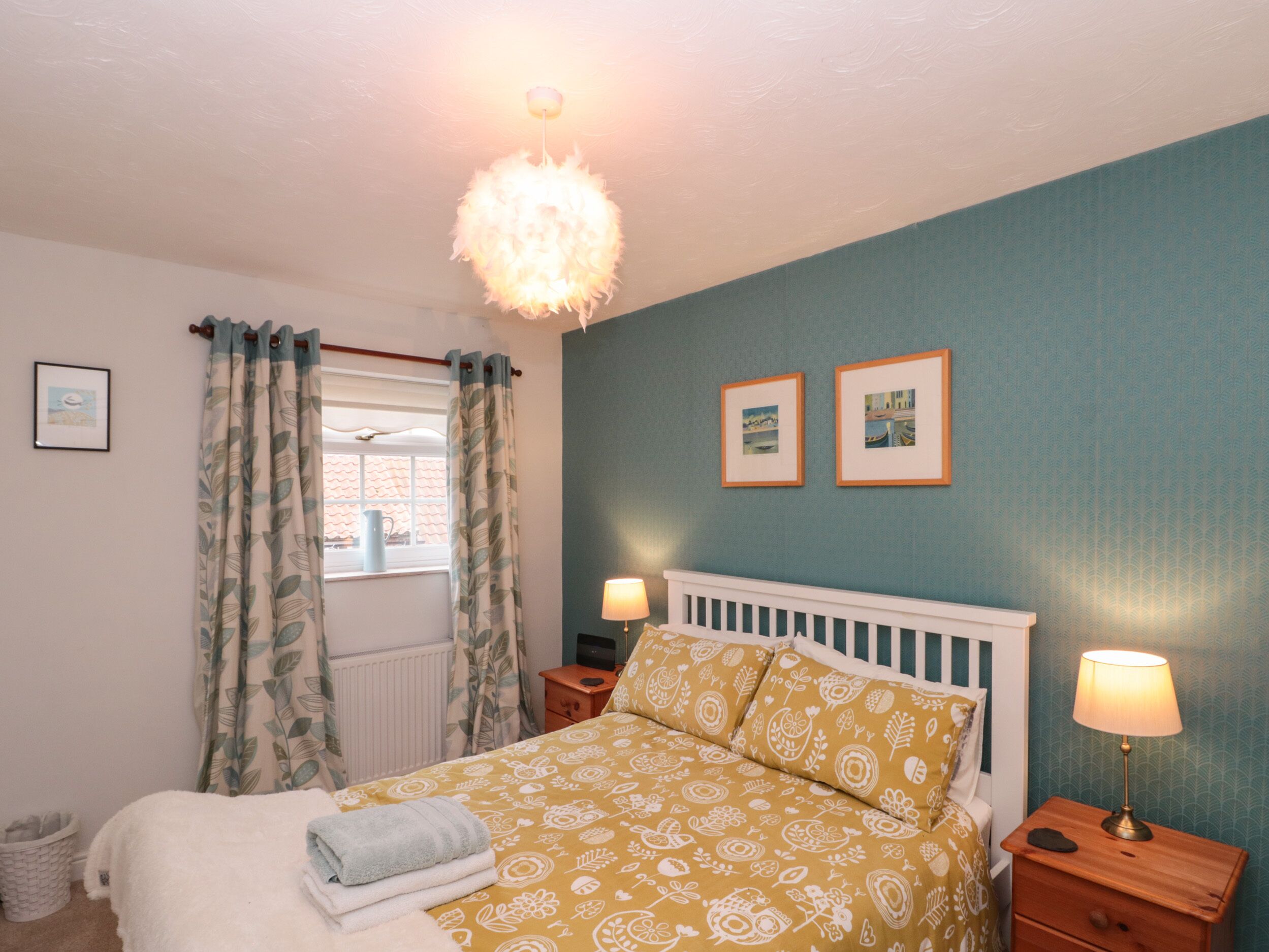 A bedroom with a bed, two bedside tables, a window with curtains, a radiator, and two framed pictures on the wall.