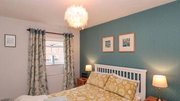 A bedroom with a bed, two bedside tables, a window with curtains, a radiator, and two framed pictures on the wall.