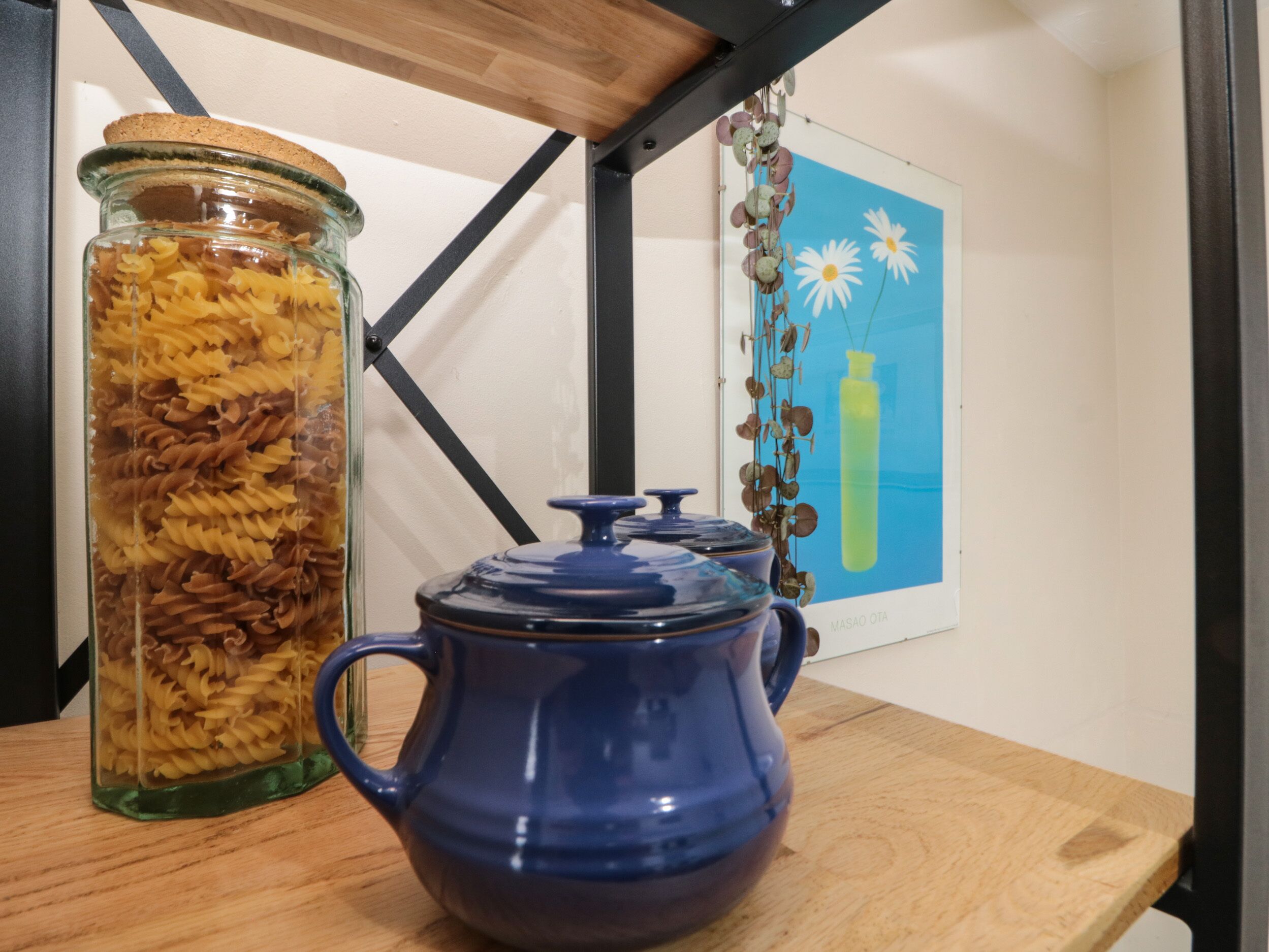 A kitchen counter with a glass jar of pasta, a blue teapot, and a framed picture on the wall.