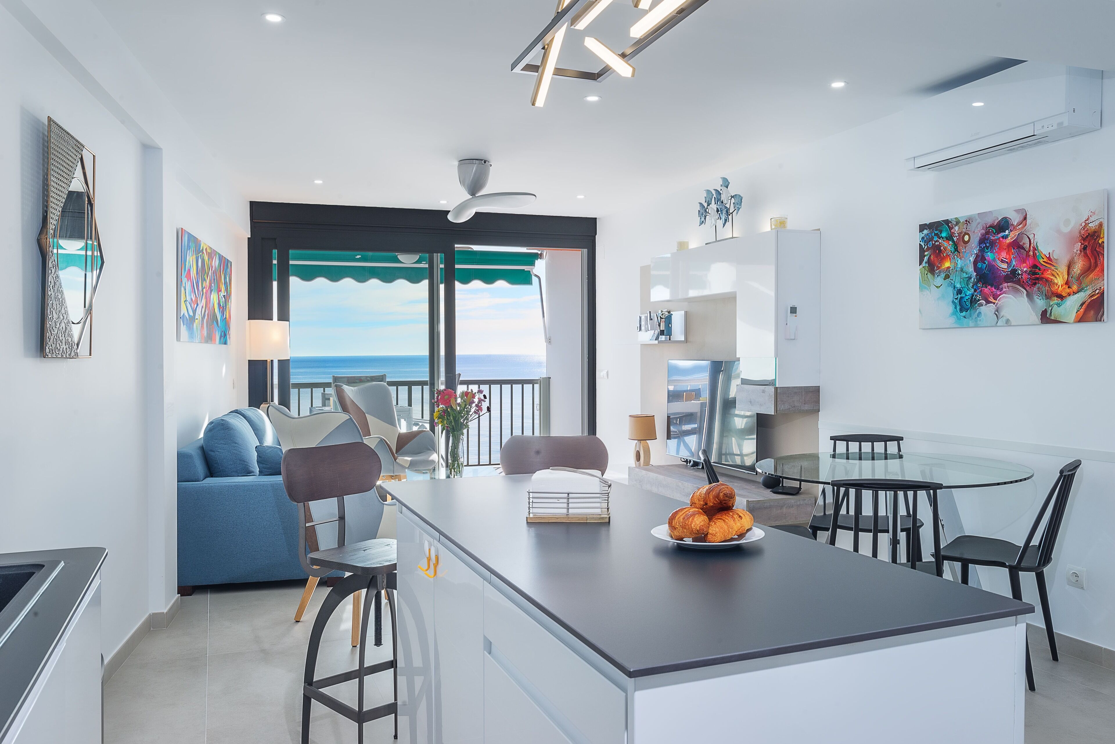 Een moderne keuken met een centraal eiland, een eethoek met een glazen tafel en uitzicht op de zee door grote ramen.